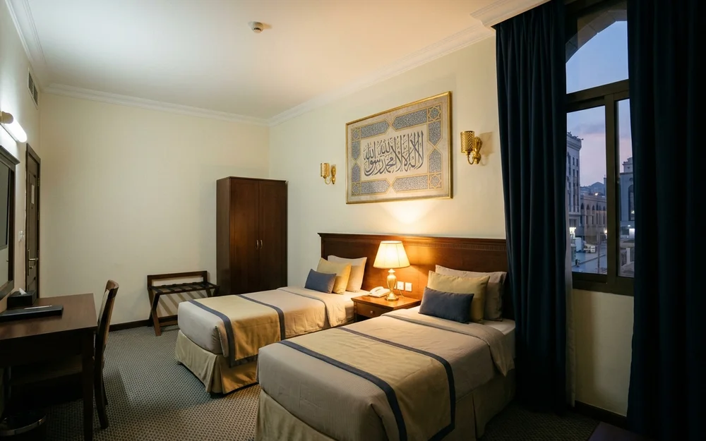 Clean three-star hotel room interior in Makkah with twin beds and warm lamp light